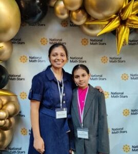 Two individuals standing in front of a "National Math Stars" backdrop decorated with gold balloons. One is an adult wearing a blue dress and a badge, and the other is a younger person in a pink sweater. Both are smiling.