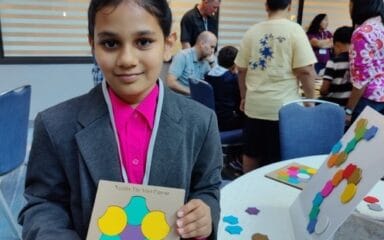 A child in a formal suit and pink shirt holds up a colorful project titled "Touch the Math Surface" at a busy event.