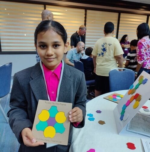 A child in a formal suit and pink shirt holds up a colorful project titled "Touch the Math Surface" at a busy event.